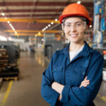 Young smiling female technician in blue uniform, protective eyeglasses and helmet working in modern factory